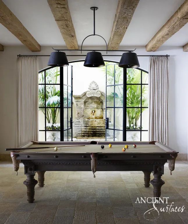 Antique limestone wall fountain crowned by a carved shell crest, framed by steel garden doors in a billiards room floored in reclaimed limestone