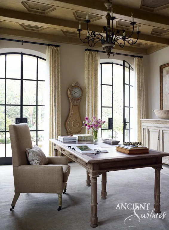 Beamed study with arched steel windows and a carved stone basin resting on the painted cabinet beyond the writing table