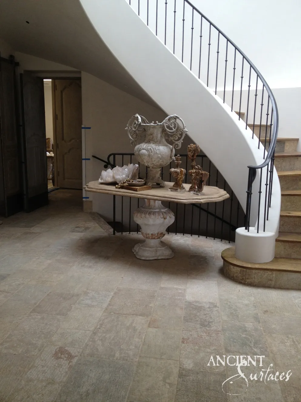 Antique limestone urn and a reclaimed limestone pedestal table set beneath a curving staircase on wide limestone floor pavers
