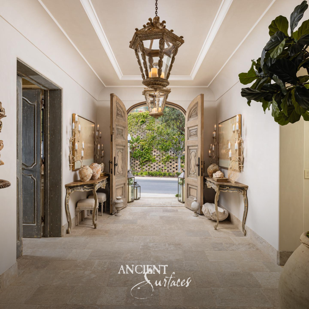 Reclaimed limestone floor pavers running the length of an entry hall between arched wooden doors and antique console tables