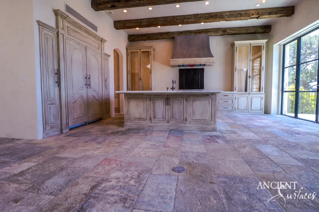 Biblical limestone kitchen flooring in wide pavers running past a carved island and range hood beneath rough hewn ceiling beams