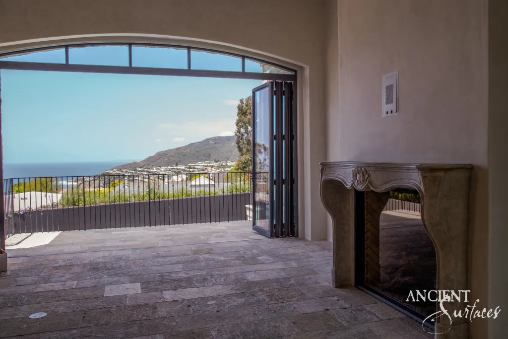 French Louis XV antique limestone fireplace mantel beside folding glass doors opening onto a hillside and ocean view